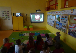 Grupa dzieci siedzi na dywanie, oglądają film edukacyjny na temat symboli narodowych.